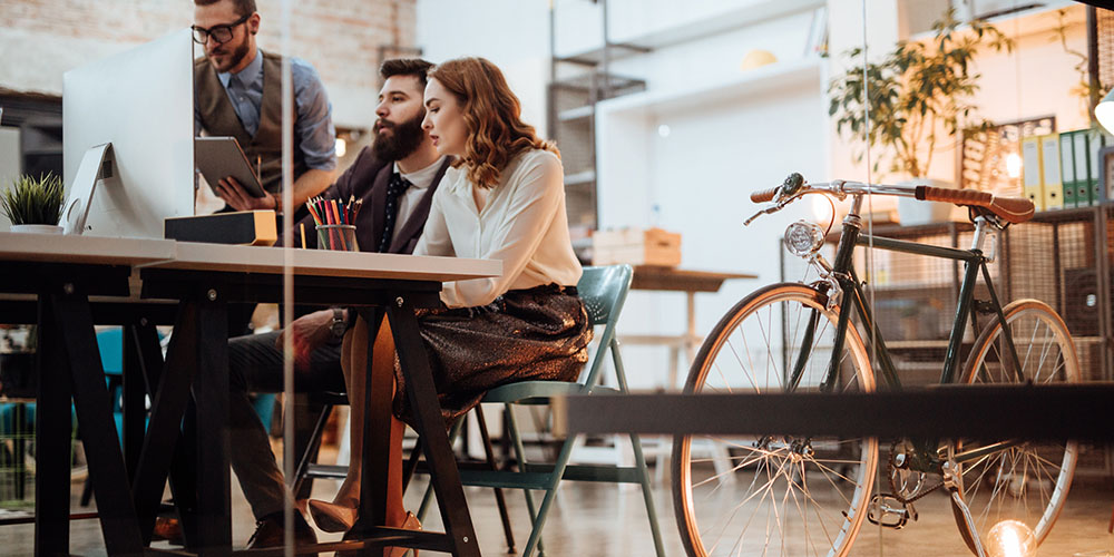 Eine Gruppe junger, hipper Menschen sitzt links im Bild an einem Tisch vor einem Laptop. Sie scheinen in geschäftliche Gespräche verwickelt. Das Umfeld spiegelt ein Büro im Industrial Style. Rechts vorne im Bild ist ein Fahrrad abgestellt.